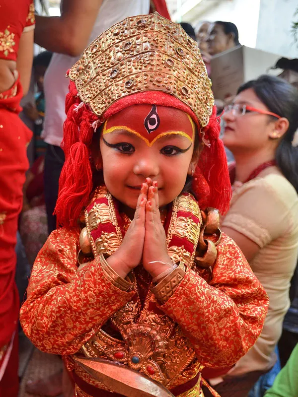 Young girl dressed as the Living Goddess Kumari in traditional red attire and golden crown offering Namaste greeting