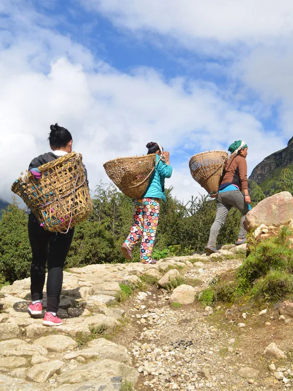 Three female local porters carrying traditional doko woven baskets up a stone stair trail on a short trek in Nepal