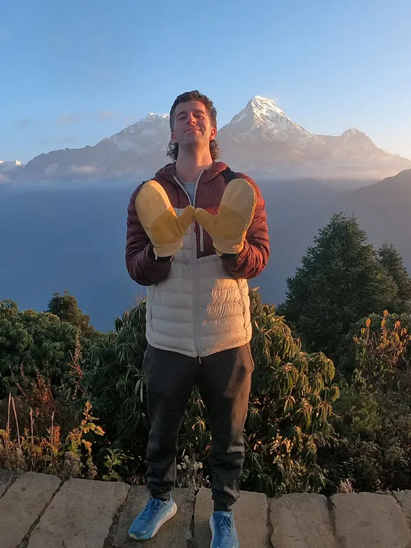 Solo male hiker posing happily in warm winter gear at Poon Hill viewpoint with Annapurna mountain range in the background.