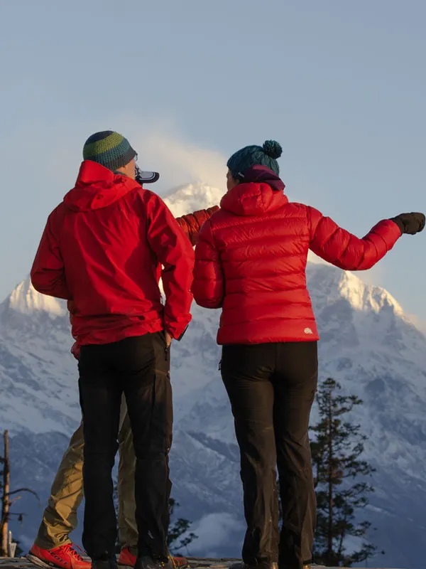 Two hikers in warm red jackets pointing toward the illuminated Himalayan snow peaks at sunrise