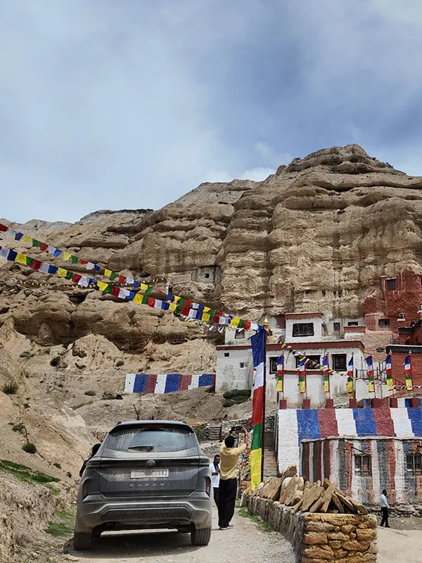 Black SUV parked on a dirt path in front of a cliffside Buddhist monastery decorated with prayer flags in Upper Mustang.