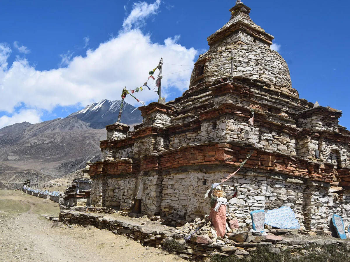 An ancient stone Buddhist chorten decorated with prayer flags along a dirt trail in Nar Phu Valley, backed by snow-dusted Himalayan mountains.