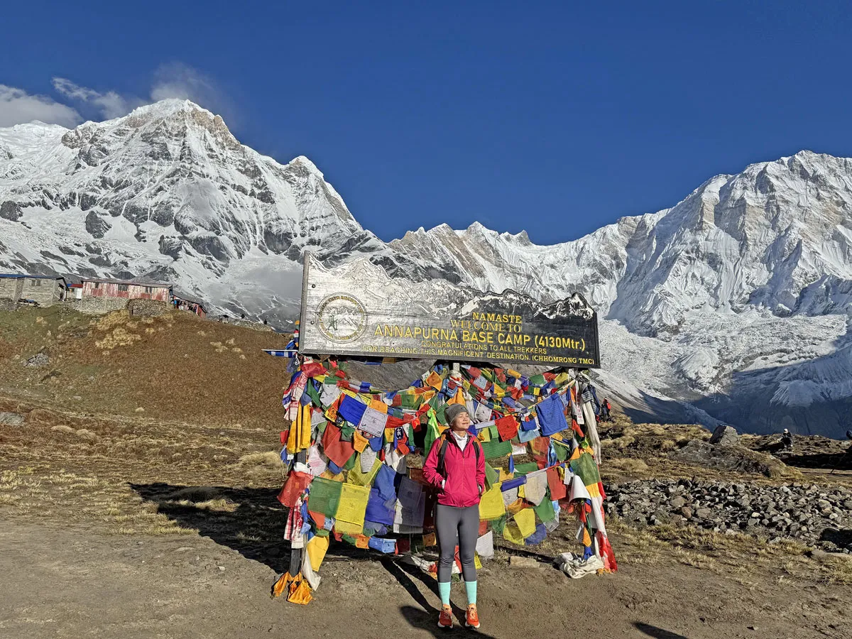 A solo trekker standing proudly in front of the Annapurna Base Camp signboard draped with colorful prayer flags and high mountain ranges behind.