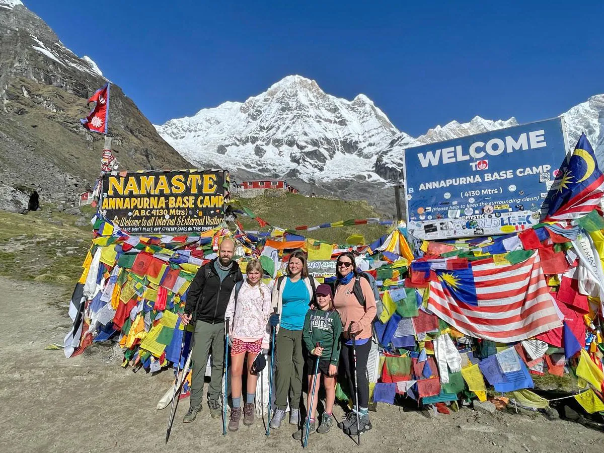 A family of trekkers posing in front of the Annapurna Base Camp welcome sign surrounded by prayer flags