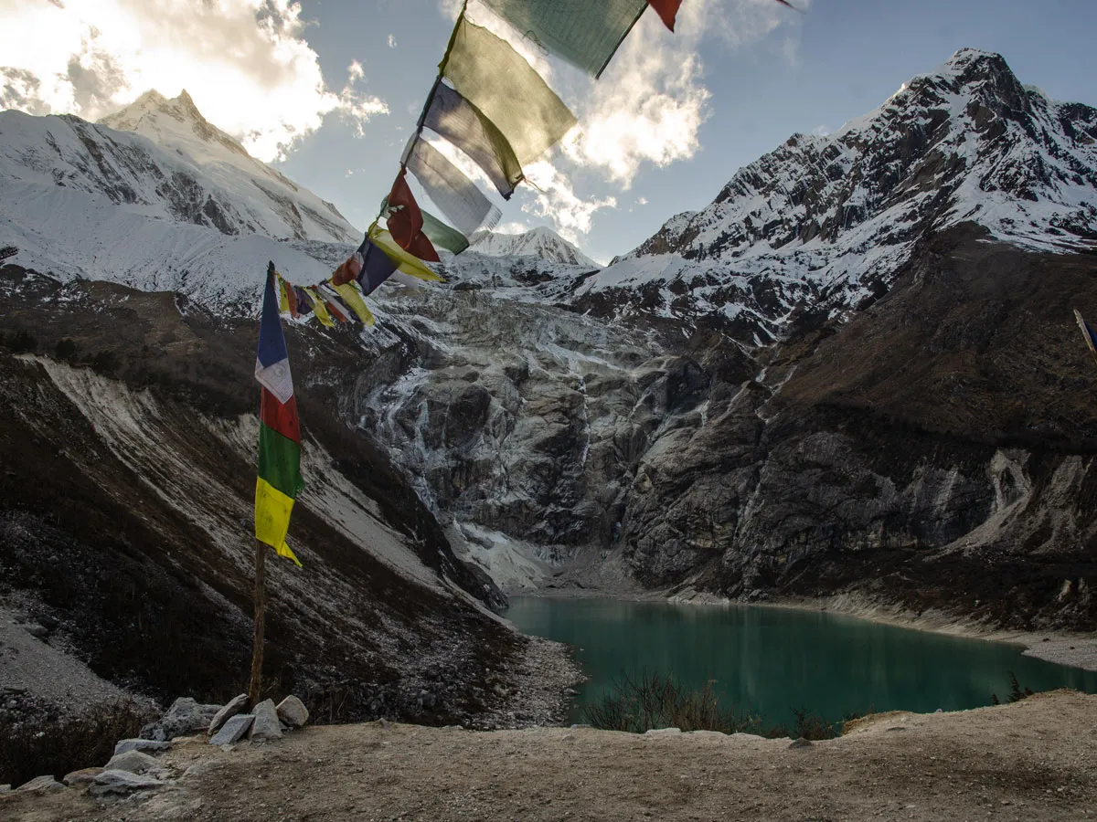 Turquoise Birendra Lake beneath steep rocky slopes and snow-covered peaks, framed by colourful Tibetan prayer flags strung overhead.