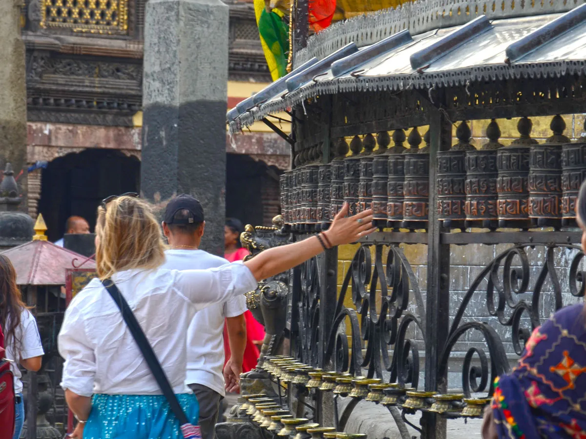 A female traveler reaching out to spin a long row of bronze Tibetan prayer wheels along the outer wall of a shrine at Boudhanath Stupa in Kathmandu, Nepal