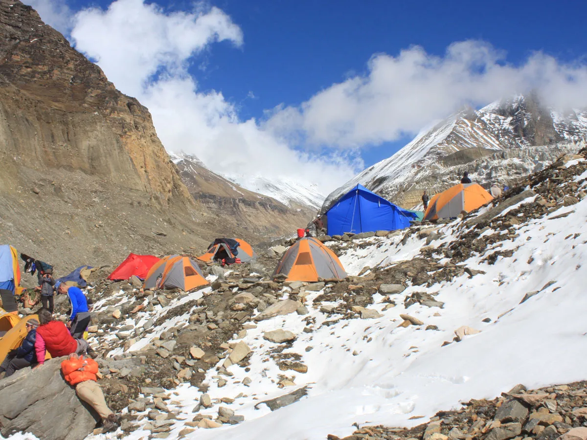Colorful expedition tents set up on rocky, snow-draped terrain surrounded by steep granite walls and glacier fields