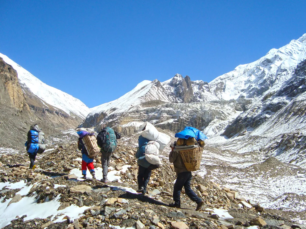 Porters carrying heavy expedition gear in traditional doko baskets along a rocky, snow-dusted glacier path