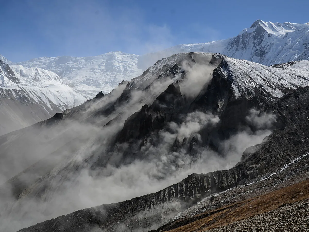 Clouds and mist rolling over dark rocky cliffs beneath massive snow-covered peaks along the Tilicho Lake trekking route in Nepal.
