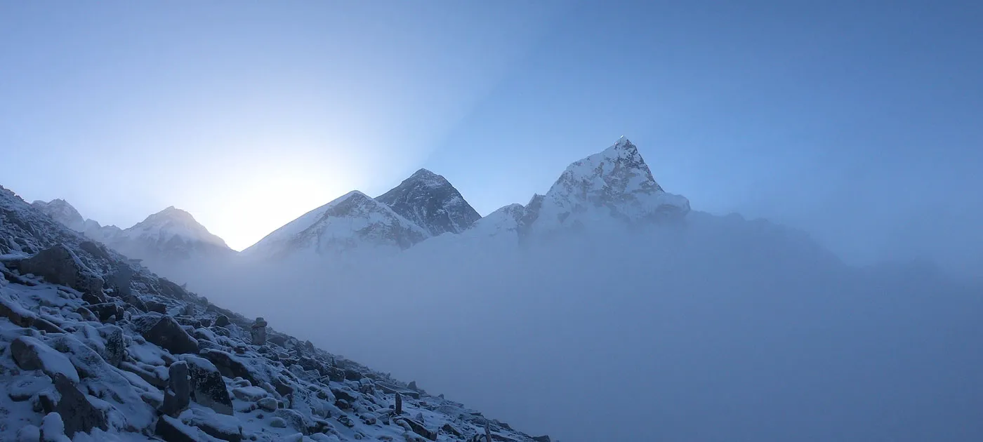 Snow-capped Himalayan peaks, including Mount Everest, emerging through morning clouds and fog above a rocky mountain slope.