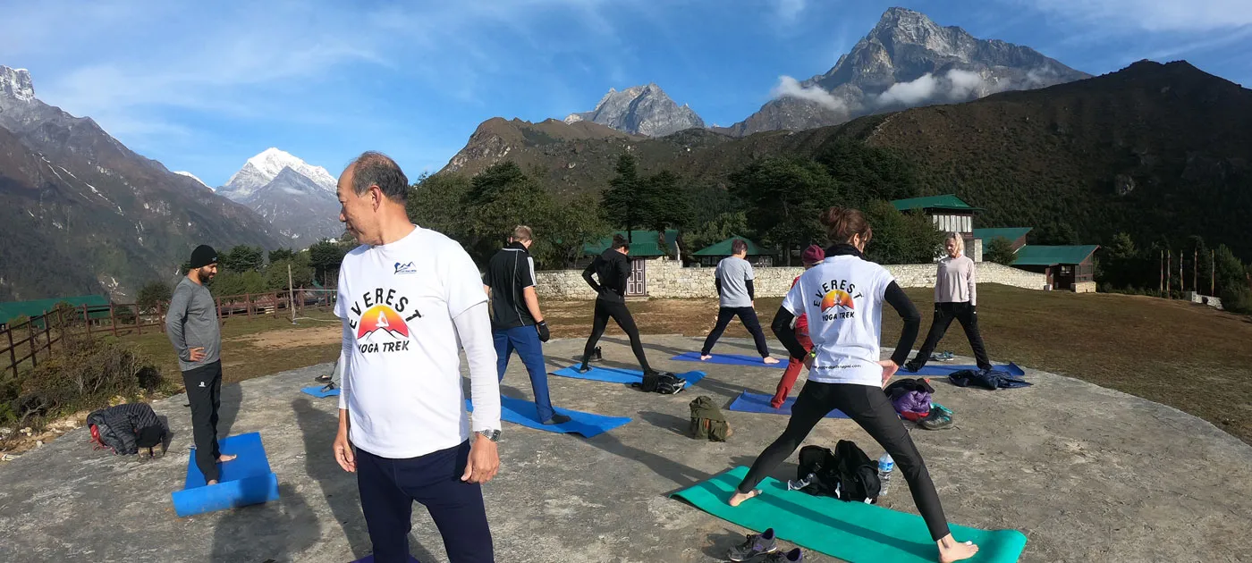 A group of trekkers wearing Everest Yoga Trek long-sleeve shirts practicing Warrior pose on blue and green yoga mats on a stone terrace