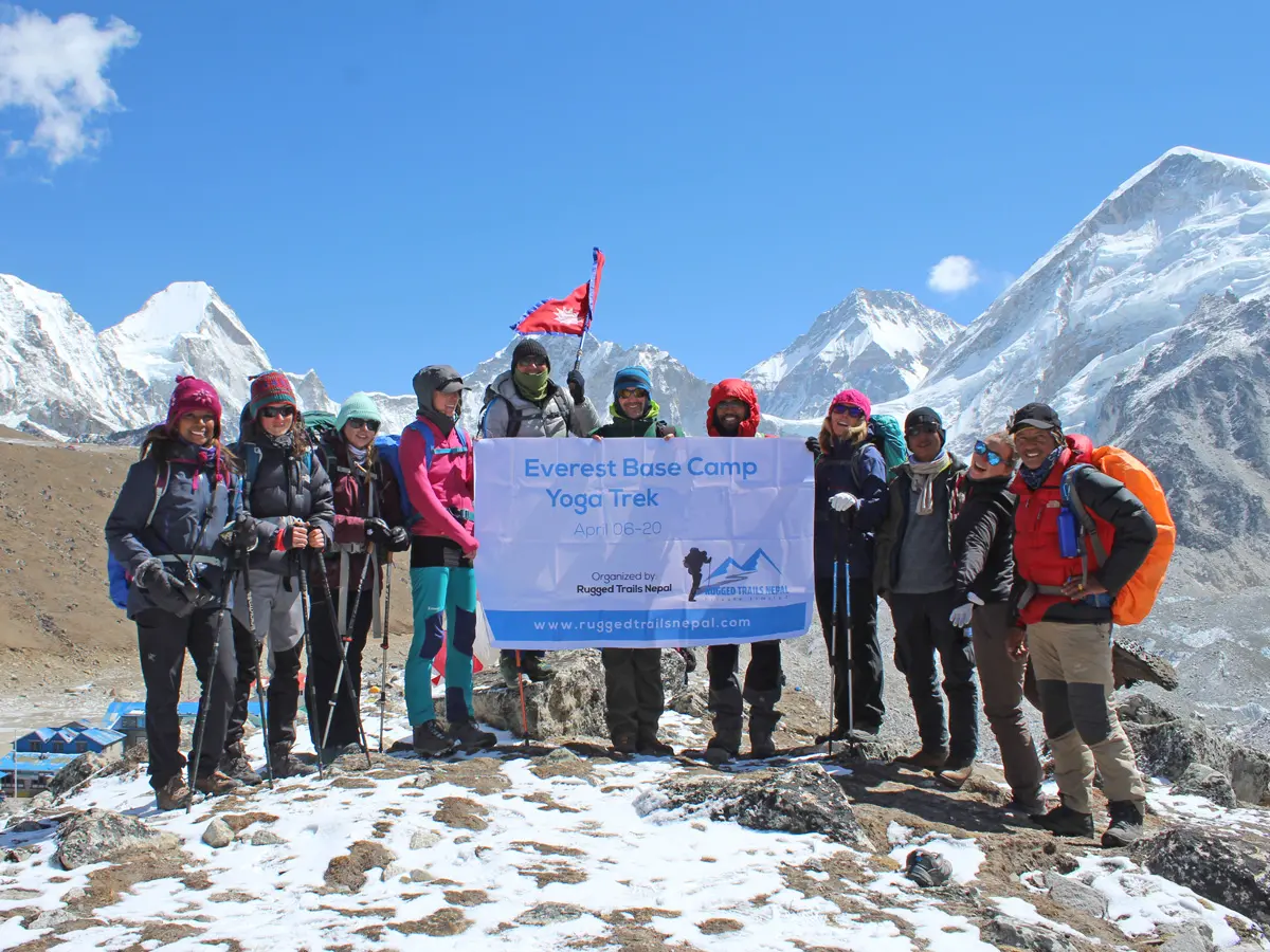 A group of happy trekkers holding the Everest Base Camp Yoga Trek banner on a snowy mountain ridge.
