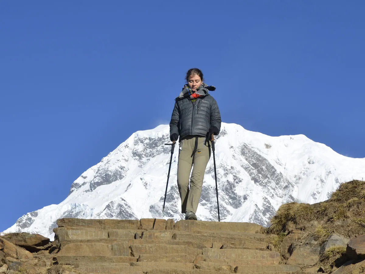 Female hiker with trekking poles descending wide stone stairs with a massive snow-covered Himalayan mountain directly behind her.