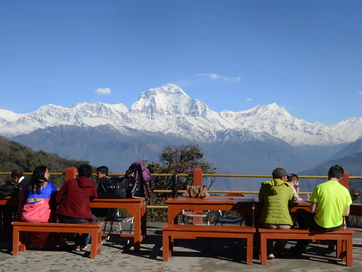 Trekkers relaxing on wooden benches at a mountain teahouse enjoying views of Mount Dhaulagiri and the Annapurna range in Nepal.
