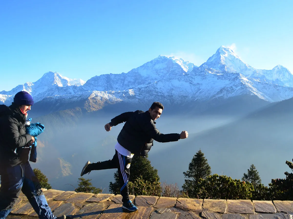 A male trekker posing in a running action mid-air on a stone path at Poon Hill, with snow-capped Annapurna peaks rising behind him under a clear blue sky.