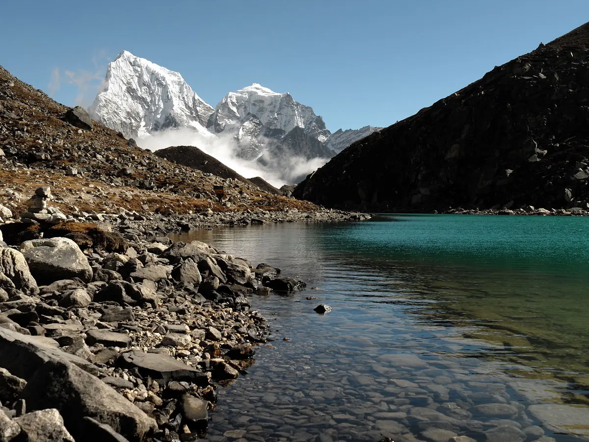 Clear turquoise waters of a Gokyo Lake with rocky shoreline and snow-capped peaks in the background.