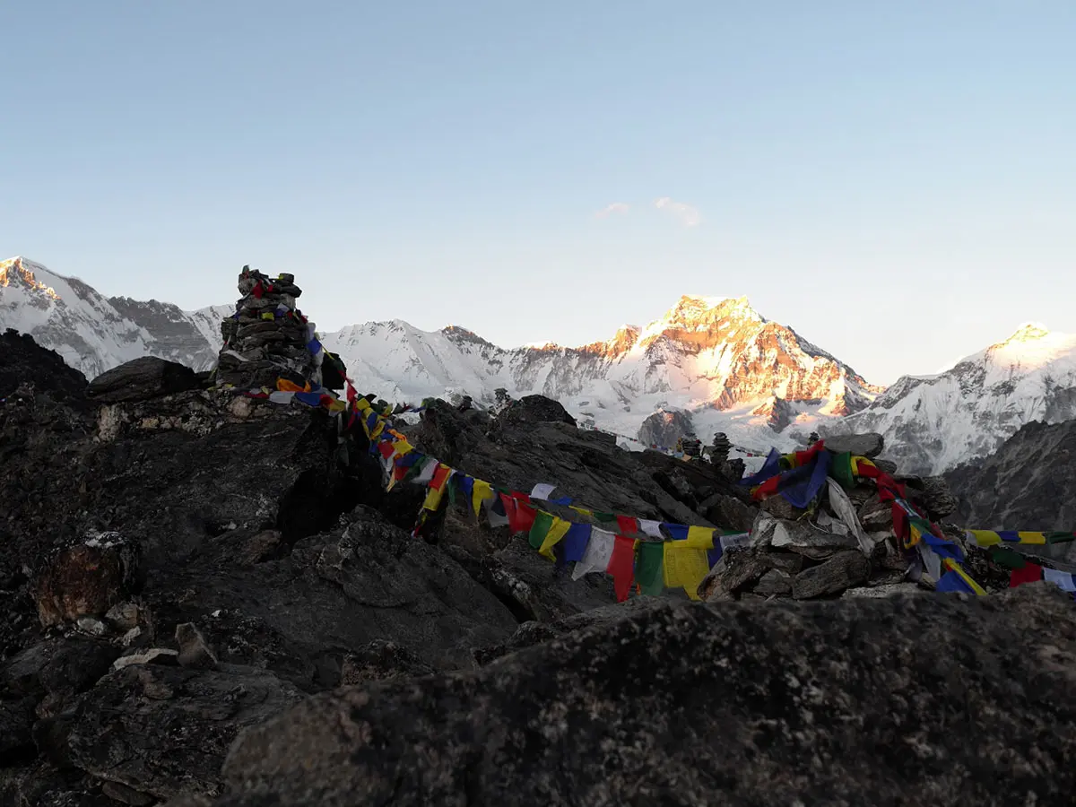 Buddhist prayer flags and rock cairns on a rocky mountain ridge with golden Himalayan peaks at sunrise.