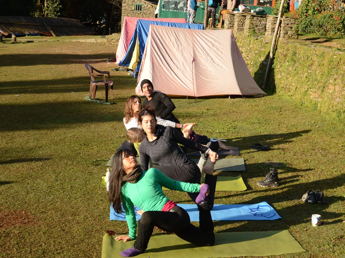 A group of trekkers practicing stretching yoga poses together on mats in a grassy campsite setting with tents in the background.