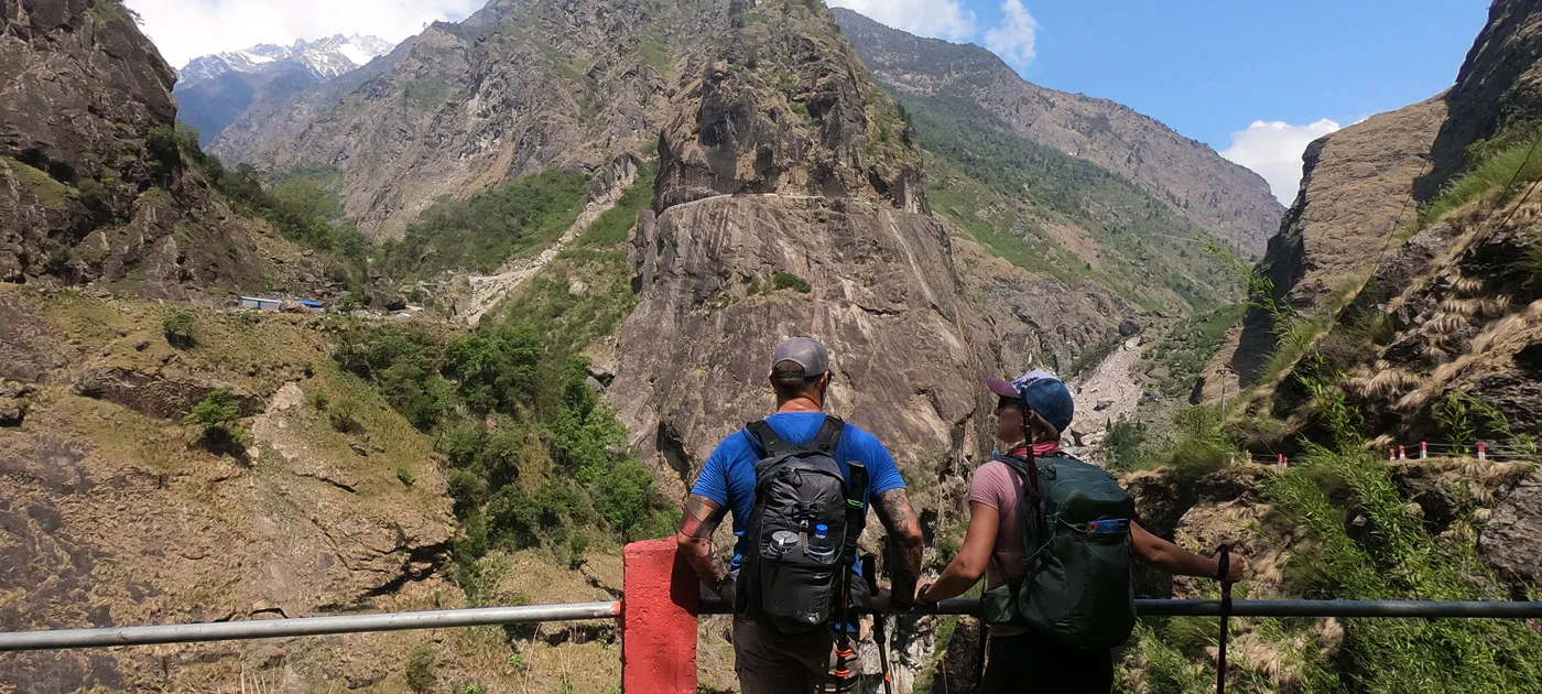 Two backpackers standing side-by-side at a railing looking at a steep mountain valley and rocky gorge in Nepal.