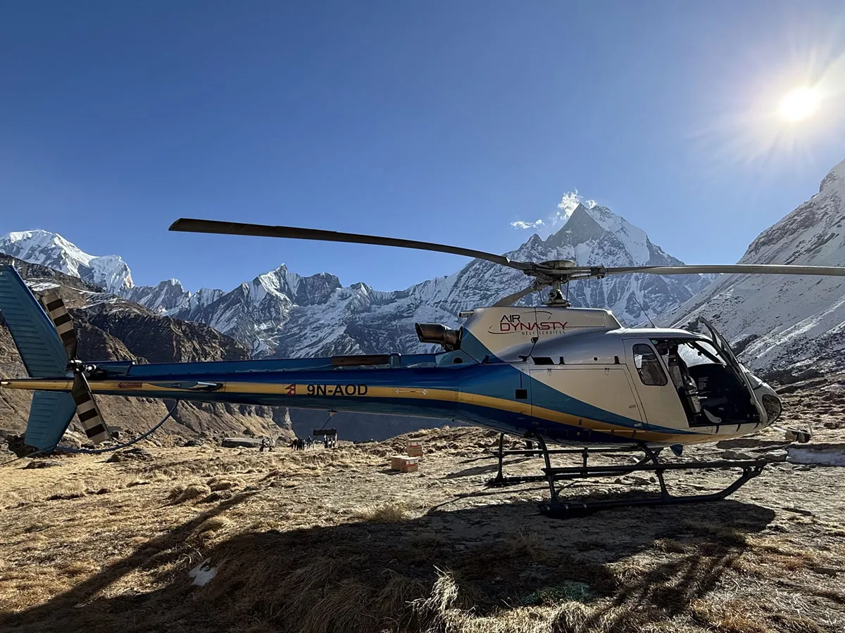 An Air Dynasty helicopter parked at Annapurna Base Camp surrounded by rugged snow-draped Himalayan mountains under a bright sunny sky