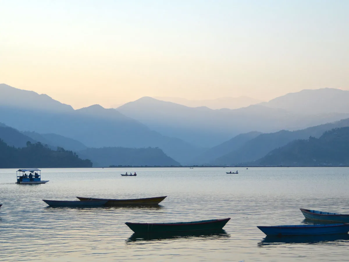 Tranquil waters of Phewa Lake in Pokhara, Nepal, featuring colorful wooden boats floating in the foreground and serene blue mountain silhouettes in the background
