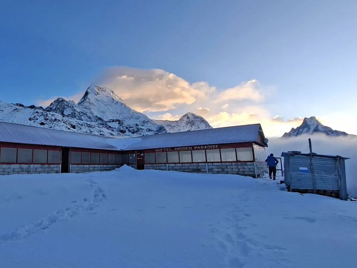 Snow-covered ground leading to Hotel Hidden Paradise teahouse with dramatic Himalayan peaks rising above the clouds.