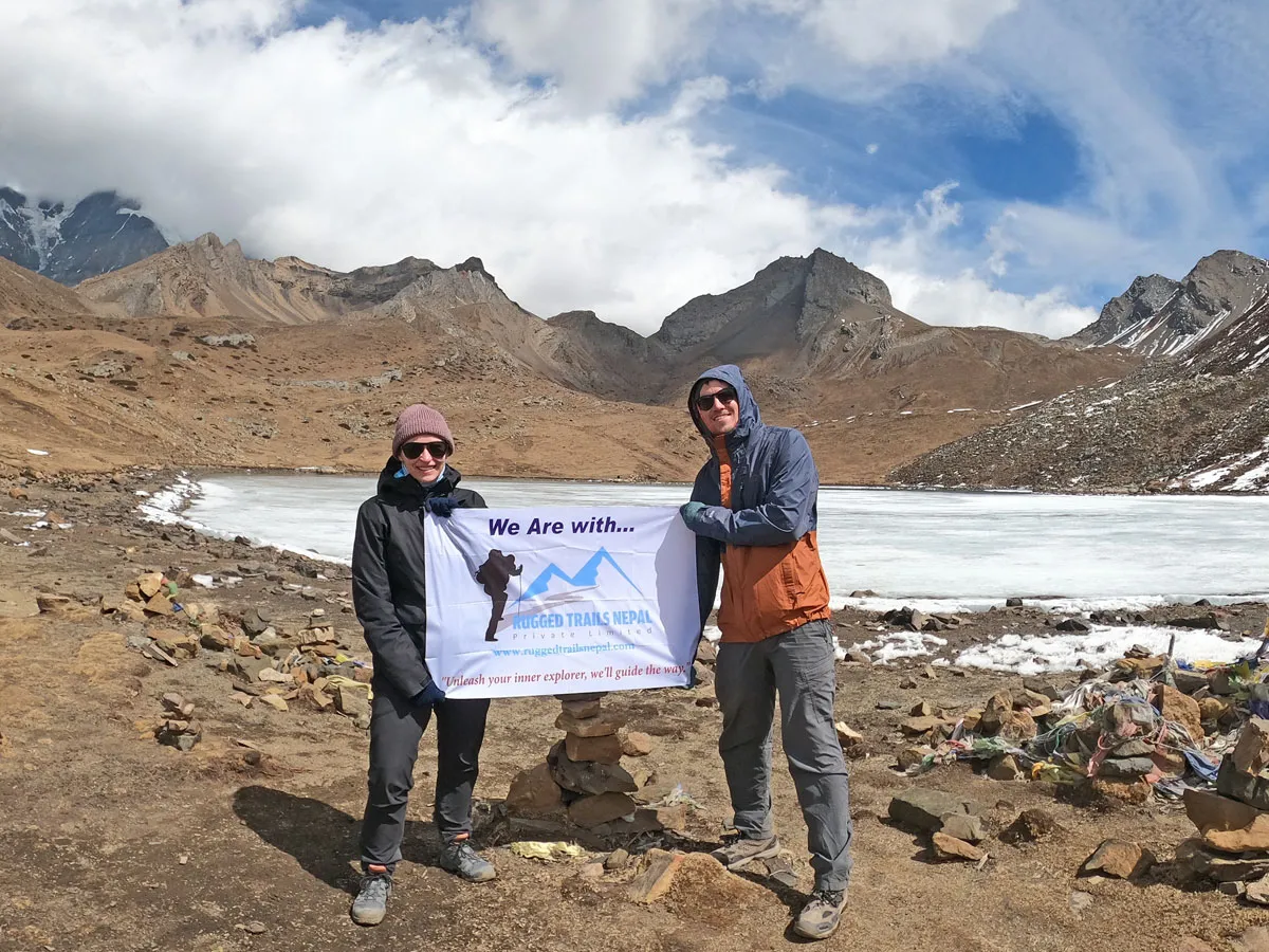 Two trekkers holding a Rugged Trails Nepal banner beside the frozen Ice Lake (Kicho Tal) in the Annapurna region.