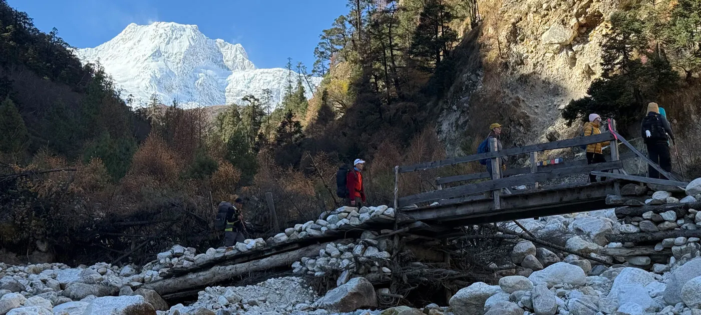 A group of foreign trekkers carrying backpacks crossing a simple wooden bridge built over a dry, rocky riverbed with snowy Himalayan mountains in the distance