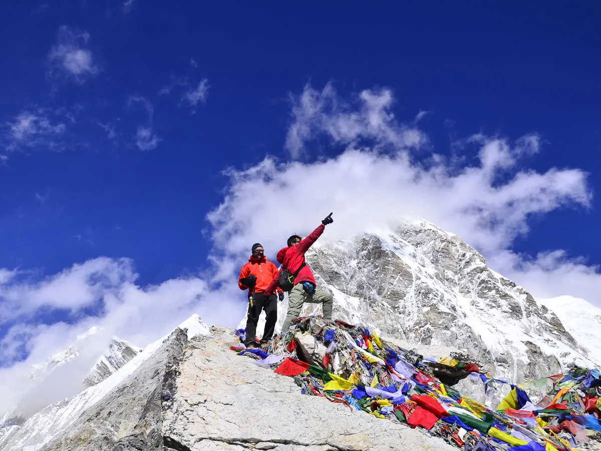 Two trekkers standing among colorful prayer flags atop a rocky summit overlooking snow-covered Himalayan peaks under a blue sky