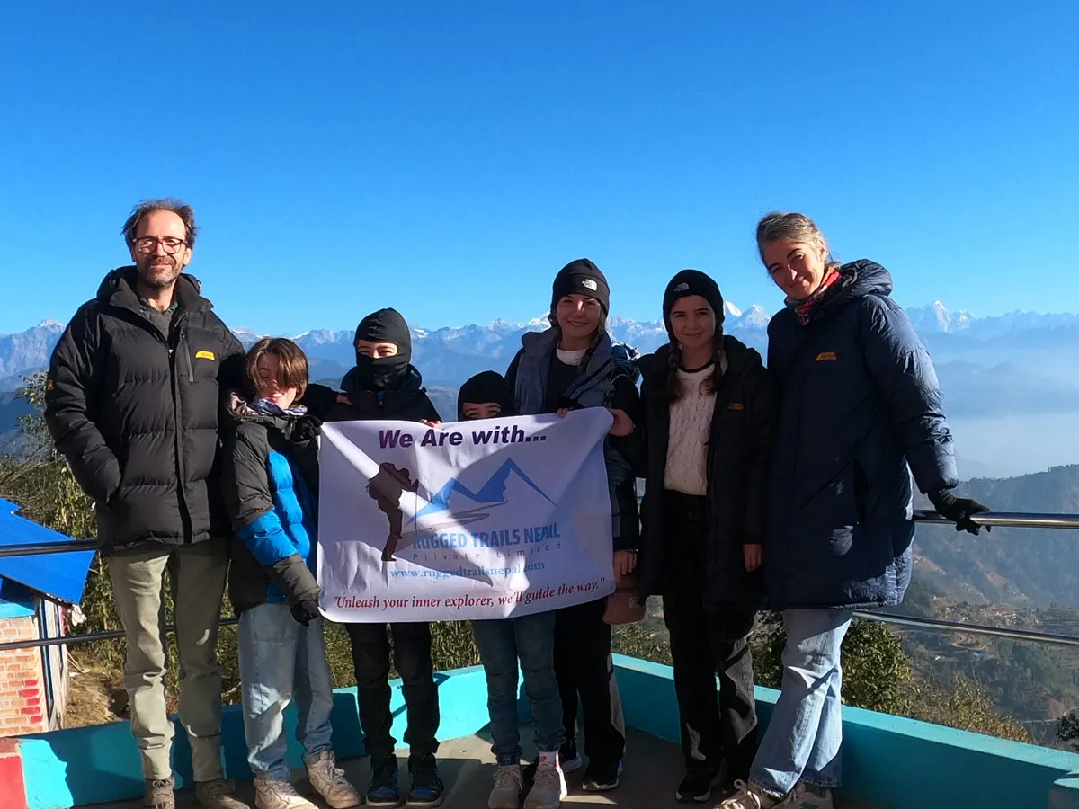 A group of seven trekkers standing together on a mountain viewpoint terrace holding a white Rugged Trails Nepal company banner against a clear sky and Himalayan backdrop