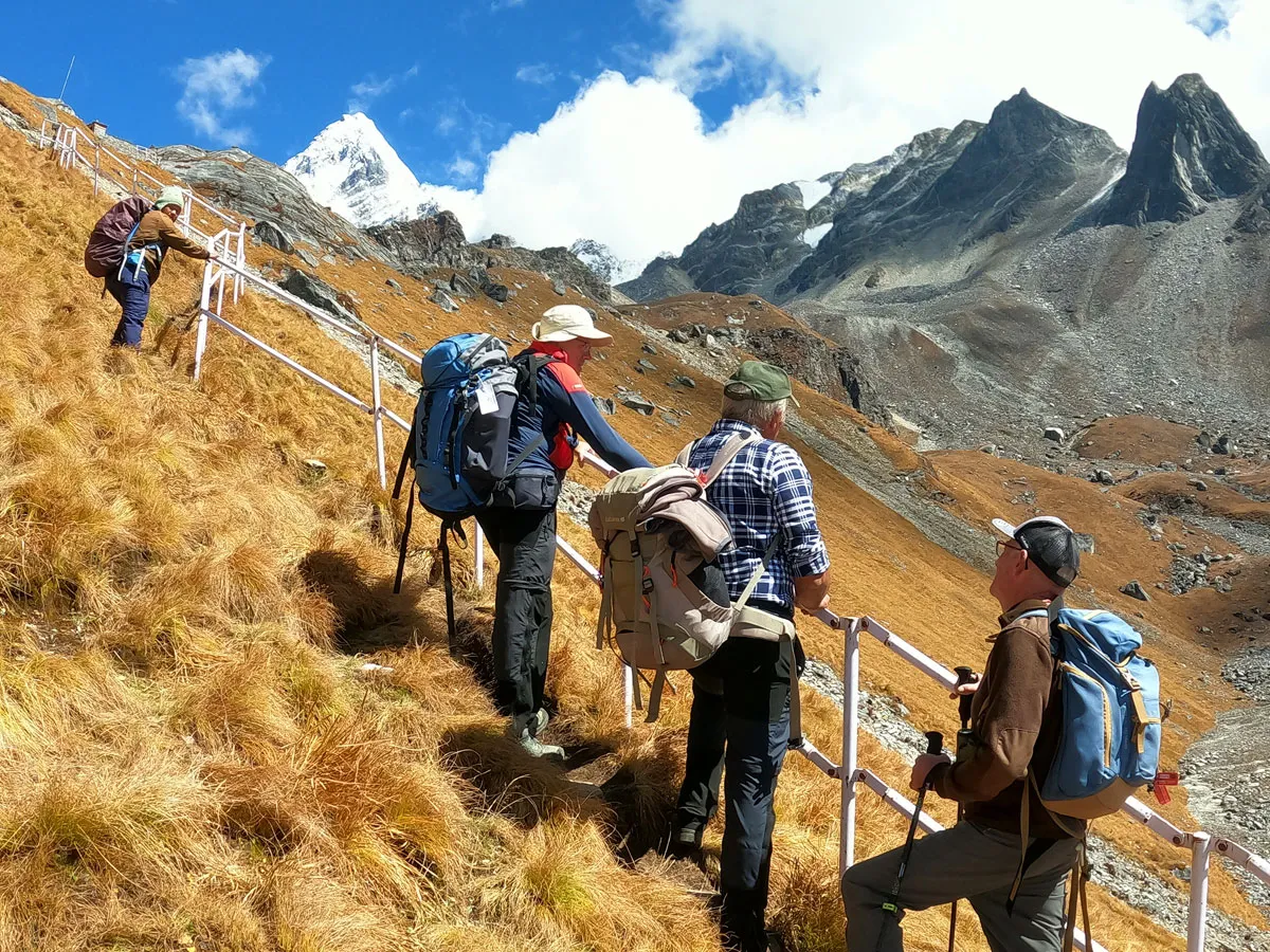 Group of trekkers with backpacks holding onto a white metal safety railing on a steep golden hill trail heading towards high rocky mountain peaks.