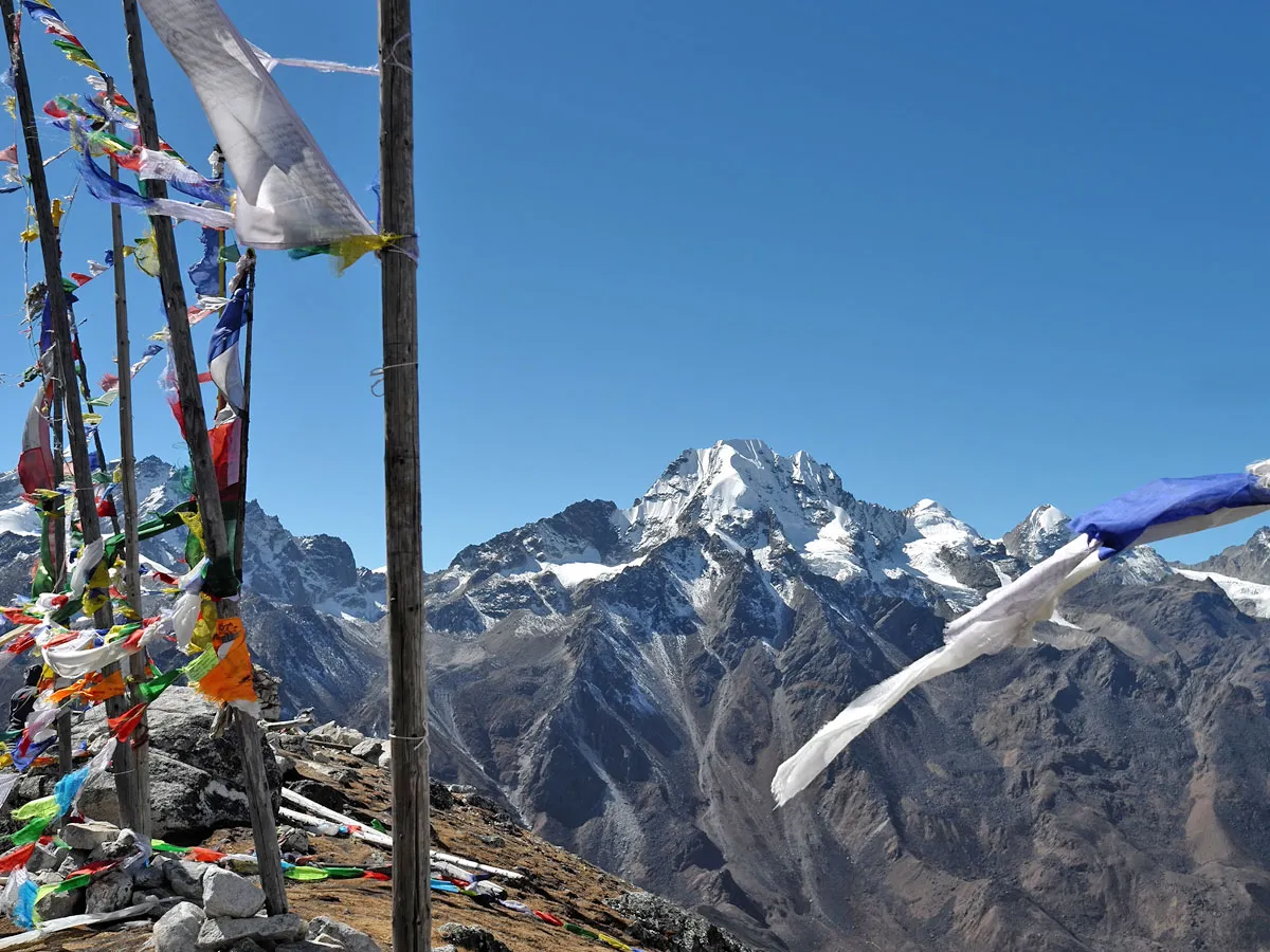 Prayer flags fluttering in the wind on a high mountain summit overlooking snow-covered Langtang Lirung peaks under a clear blue sky.