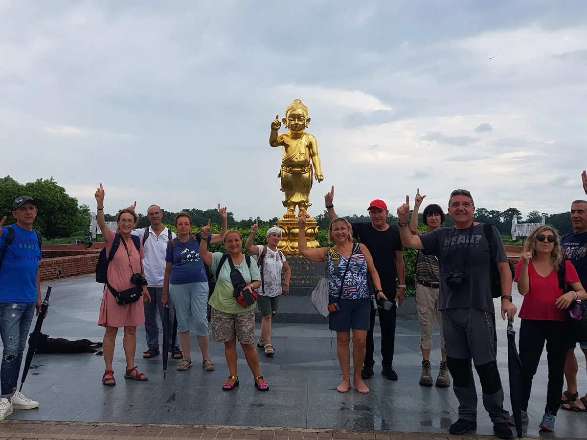 A group of international tourists posing together outdoors in front of a golden statue of Baby Buddha in Lumbini, Nepal, with many raising their index fingers in unison