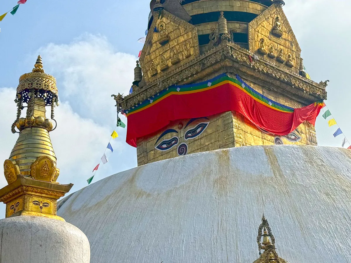 A close-up low-angle view of the gilded spire and painted eyes of Buddha at Swayambhunath Stupa in Kathmandu, draped with a bright red cloth and surrounded by prayer flags
