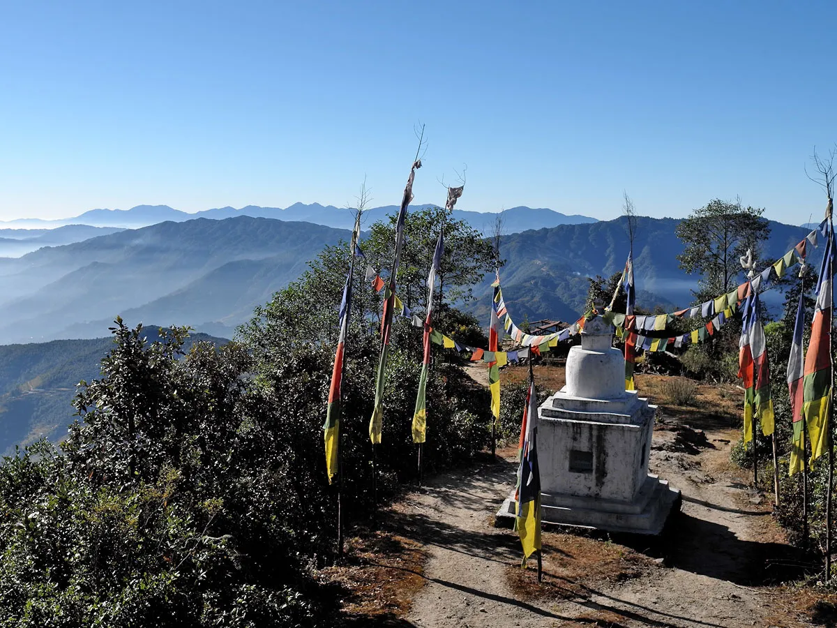 A white Buddhist chorten stupa surrounded by vertical prayer flags on a mountain ridge overlooking misty valleys on the Langtang Gosaikunda Helambu trek.
