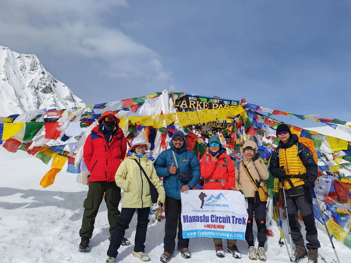 Group of six trekkers in colorful winter gear posing with a Manaslu Circuit Trek banner at Larkya La Pass amid prayer flags and snow.