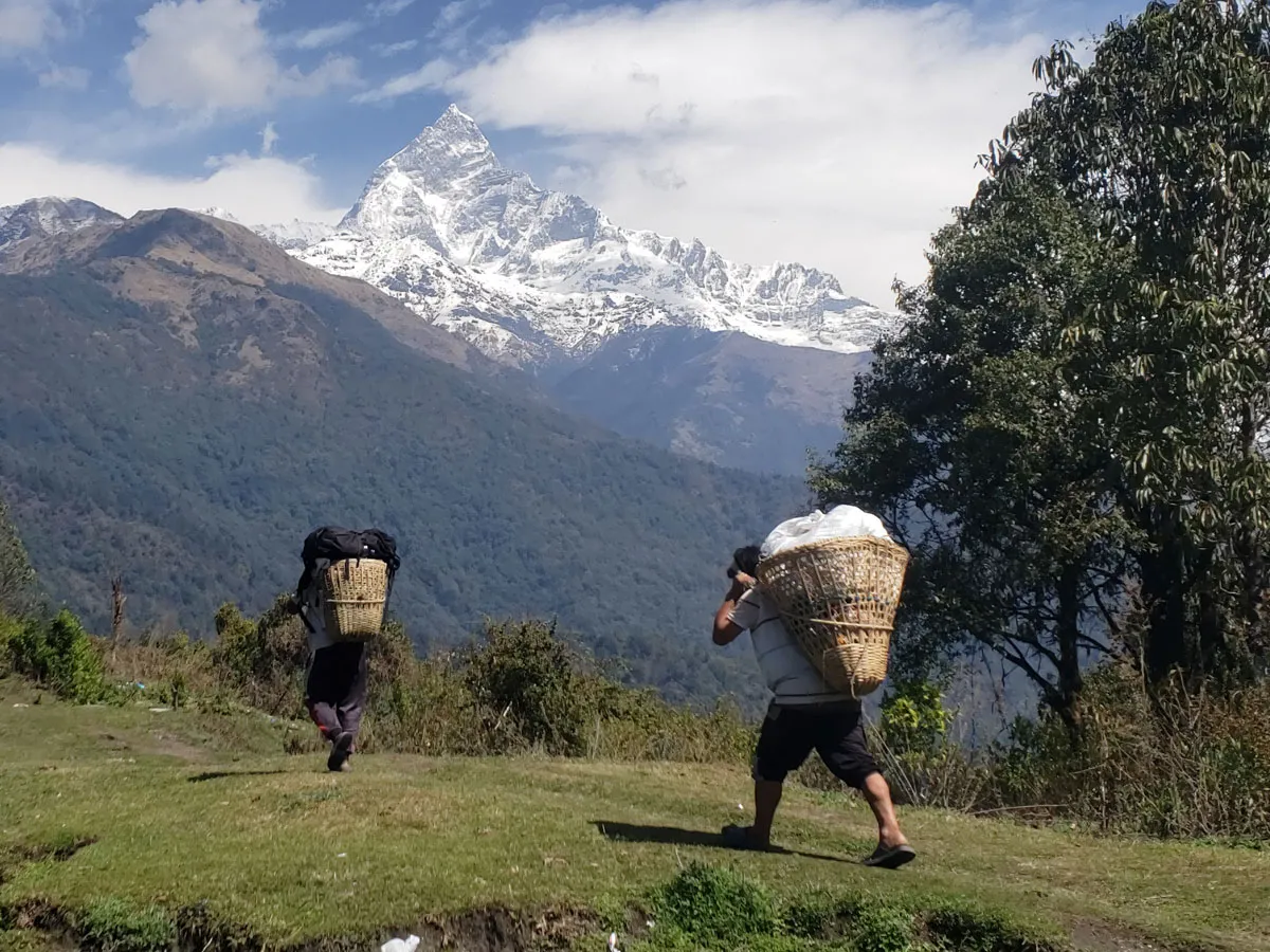 Local trekking porters carrying woven baskets with gear on a path toward Mount Machhapuchhre in the background.