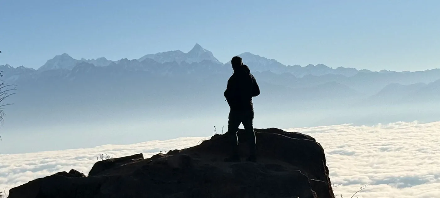 The dark silhouette of a trekker standing on a rocky ledge looking out over a thick blanket of white clouds toward distant mountain ranges