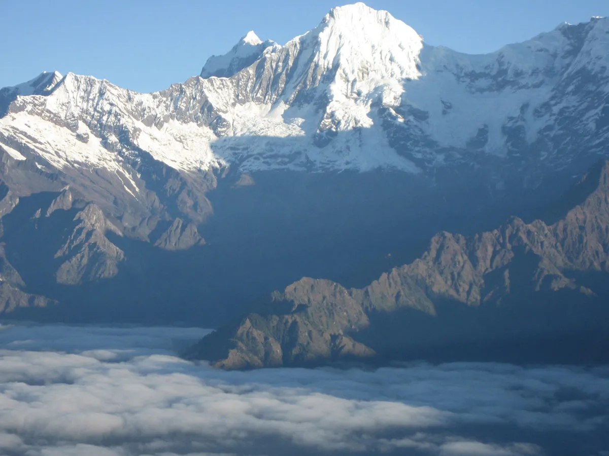 Close-up view of a high snow peak surrounded by rugged mountain slopes above a thick layer of clouds.