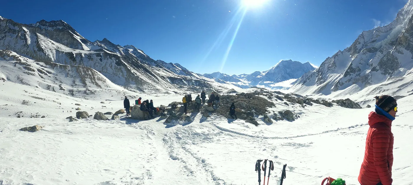 A group of trekkers resting on snow-covered ground surrounded by steep Himalayan peaks under a bright sunny sky near Larke Pass