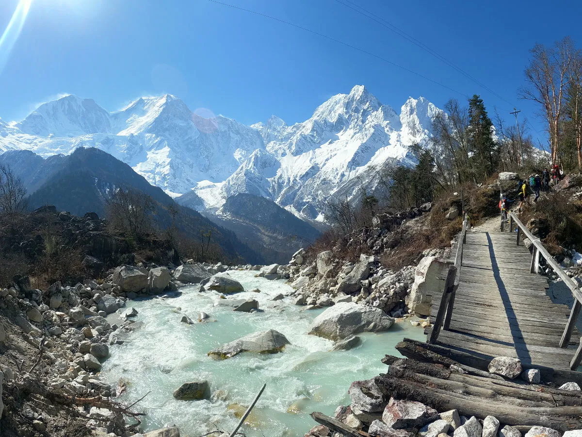 Wooden footbridge crossing a rushing turquoise mountain river with snow-capped Himalayan peaks in the background
