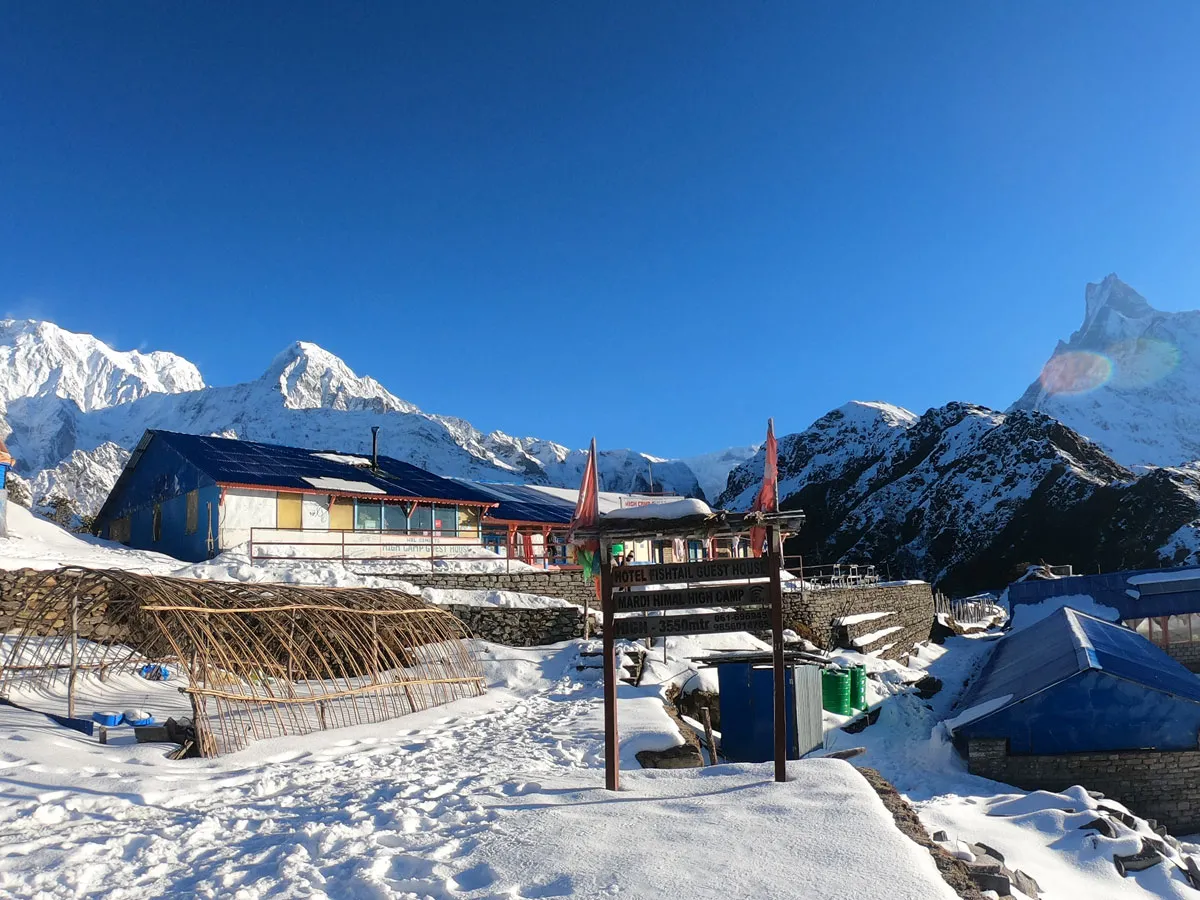 Snowy trail leading to tea houses and the Mardi Himal High Camp wooden signpost (3550m) against clear blue skies and snow-capped Himalayan peaks