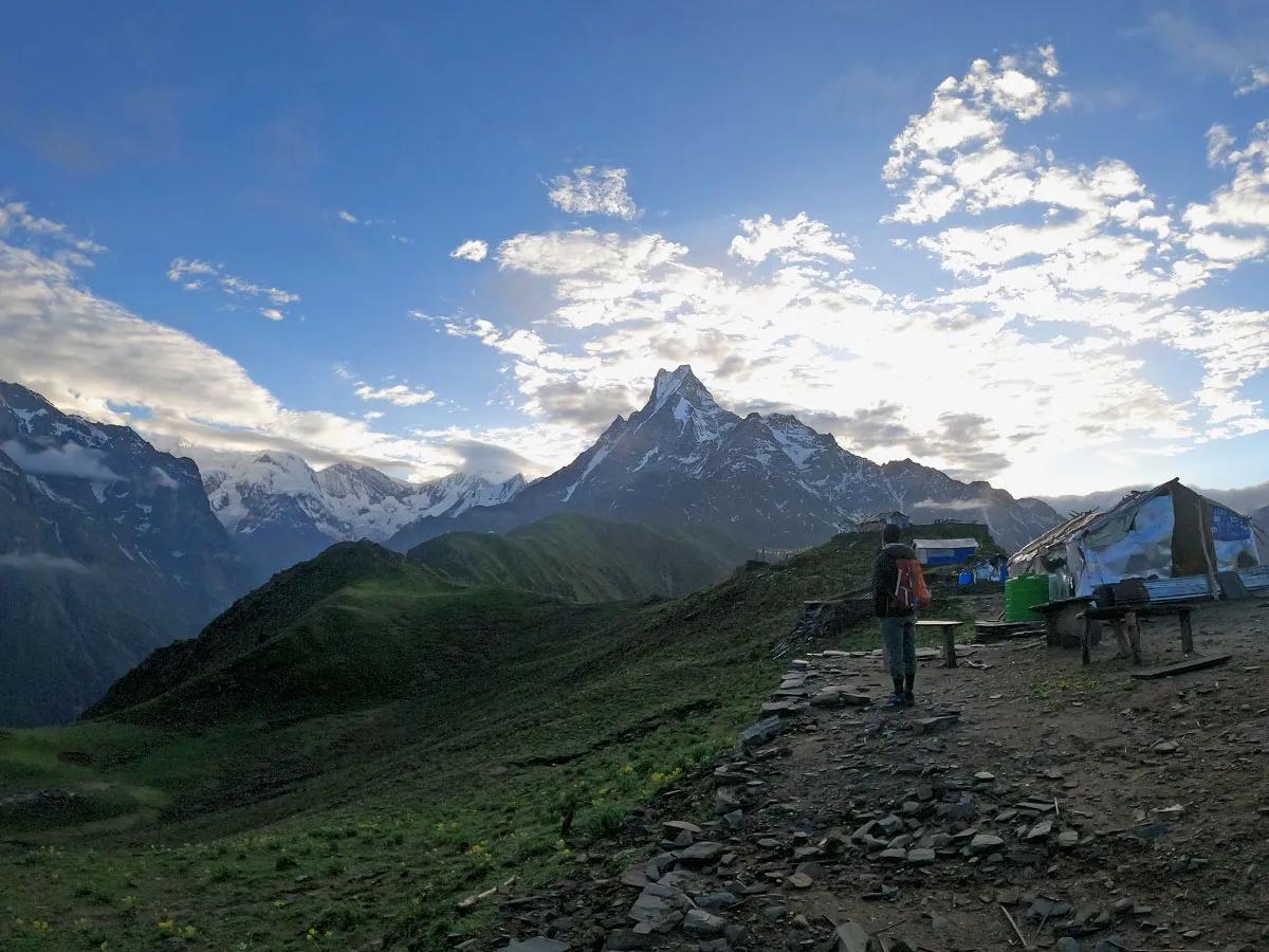 Panoramic viewpoint showing a trekker standing on a rocky ridge with Mount Machhapuchhre towering under a bright blue sky on the Mardi Himal Trek.