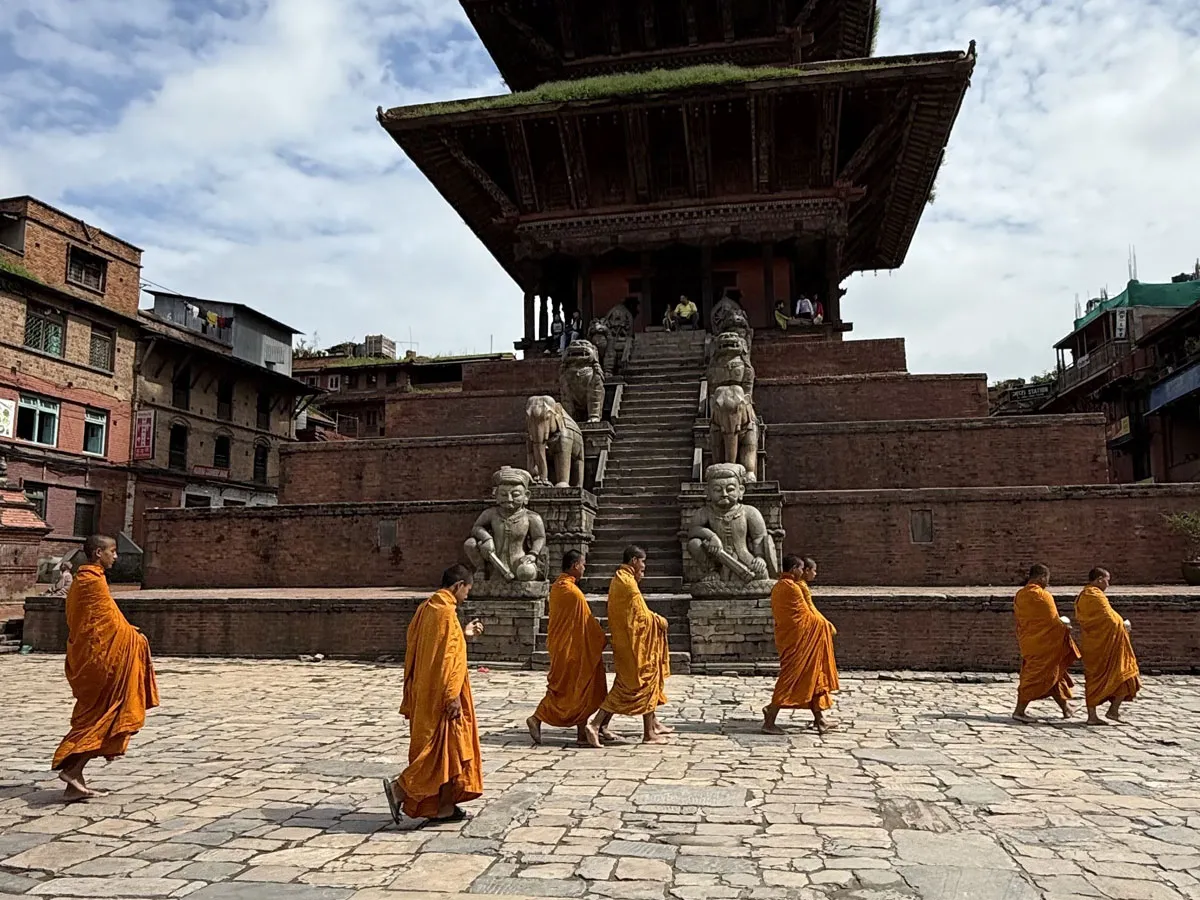 A line of Buddhist monks in traditional orange and saffron robes walking across the stone square in front of the five-tiered Nyatapola Temple in Bhaktapur