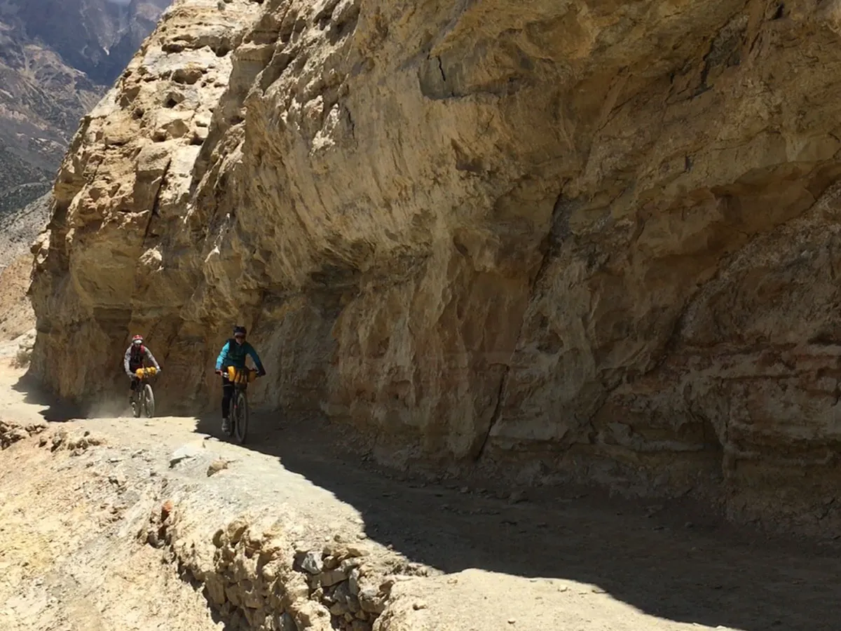 Two cyclists riding mountain bikes along a narrow dirt path directly underneath massive, overhang-eroded yellow sandstone cliffs in a desert gorge