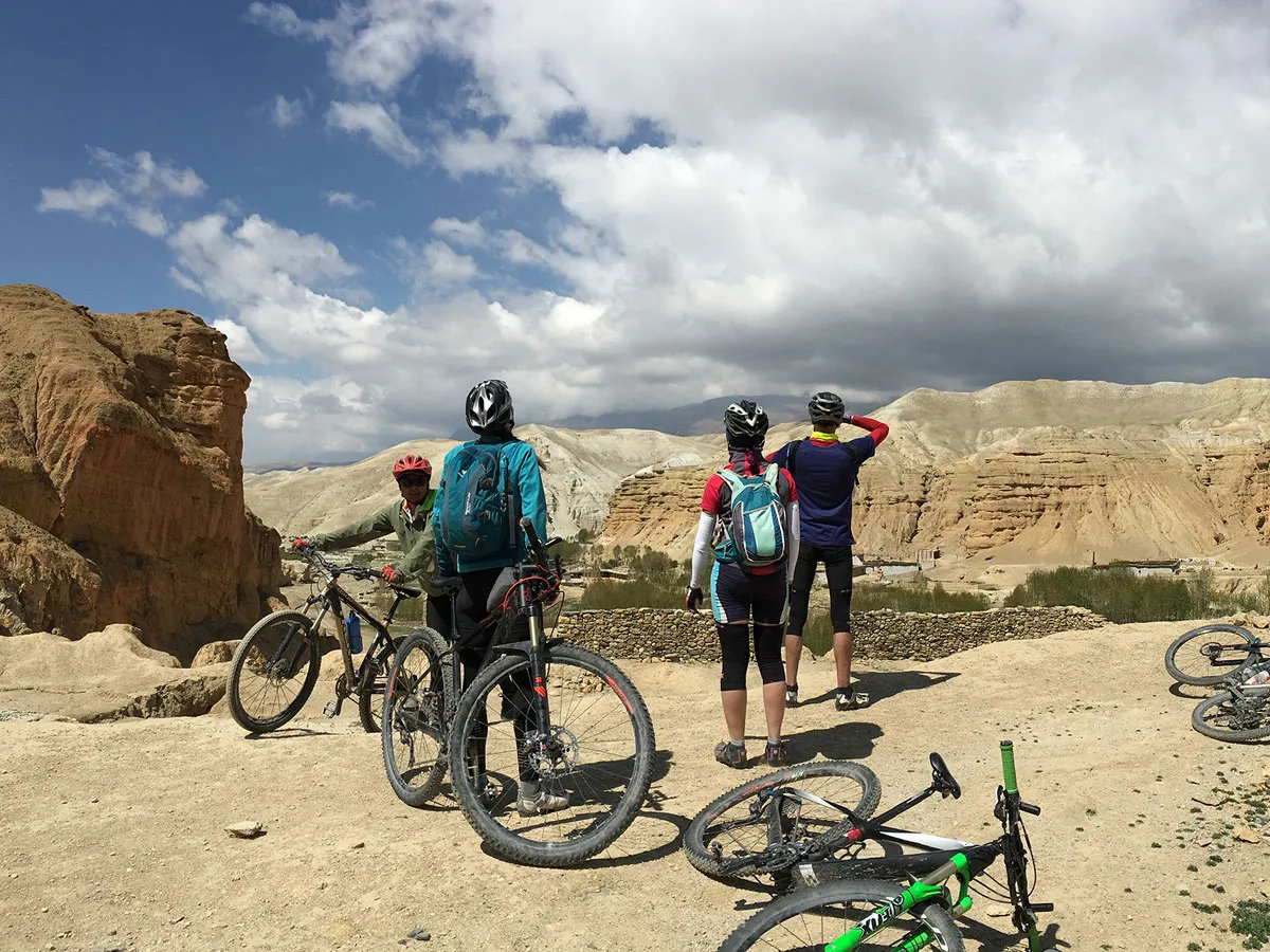 A group of four mountain bikers with helmets and backpacks standing beside their bicycles on a dusty plateau, looking out over a wide valley with unique rock formations