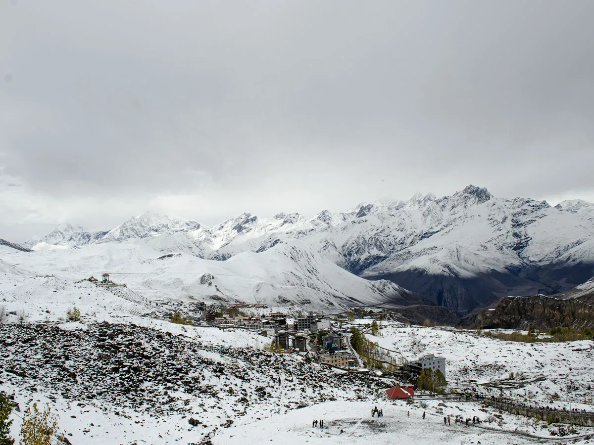 Snow-covered village street in Muktinath with local tea houses, colorful prayer flags, and dramatic snow-capped Himalayan peaks towering in the background.