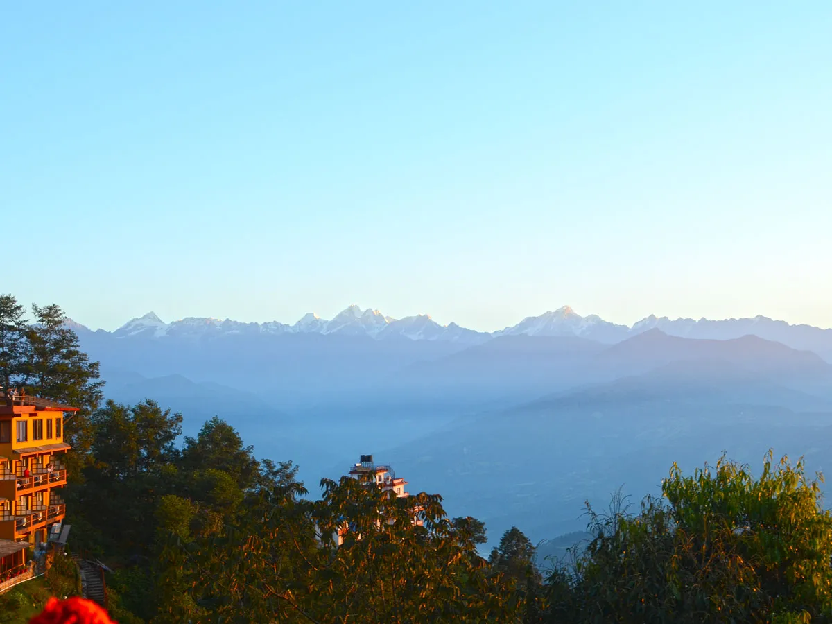Hillside resort buildings surrounded by lush trees, looking out toward the hazy Himalayan mountains