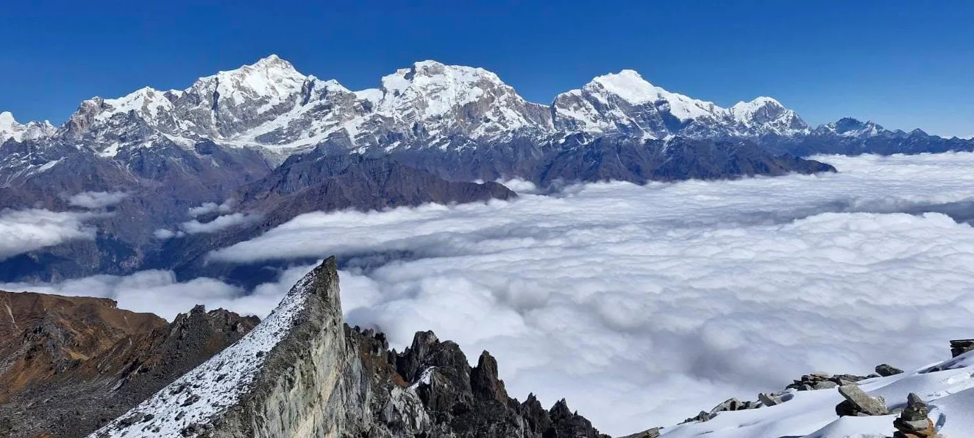 A high-altitude panoramic view looking out over a thick sea of white clouds toward majestic snow-capped Himalayan peaks under a clear blue sky.