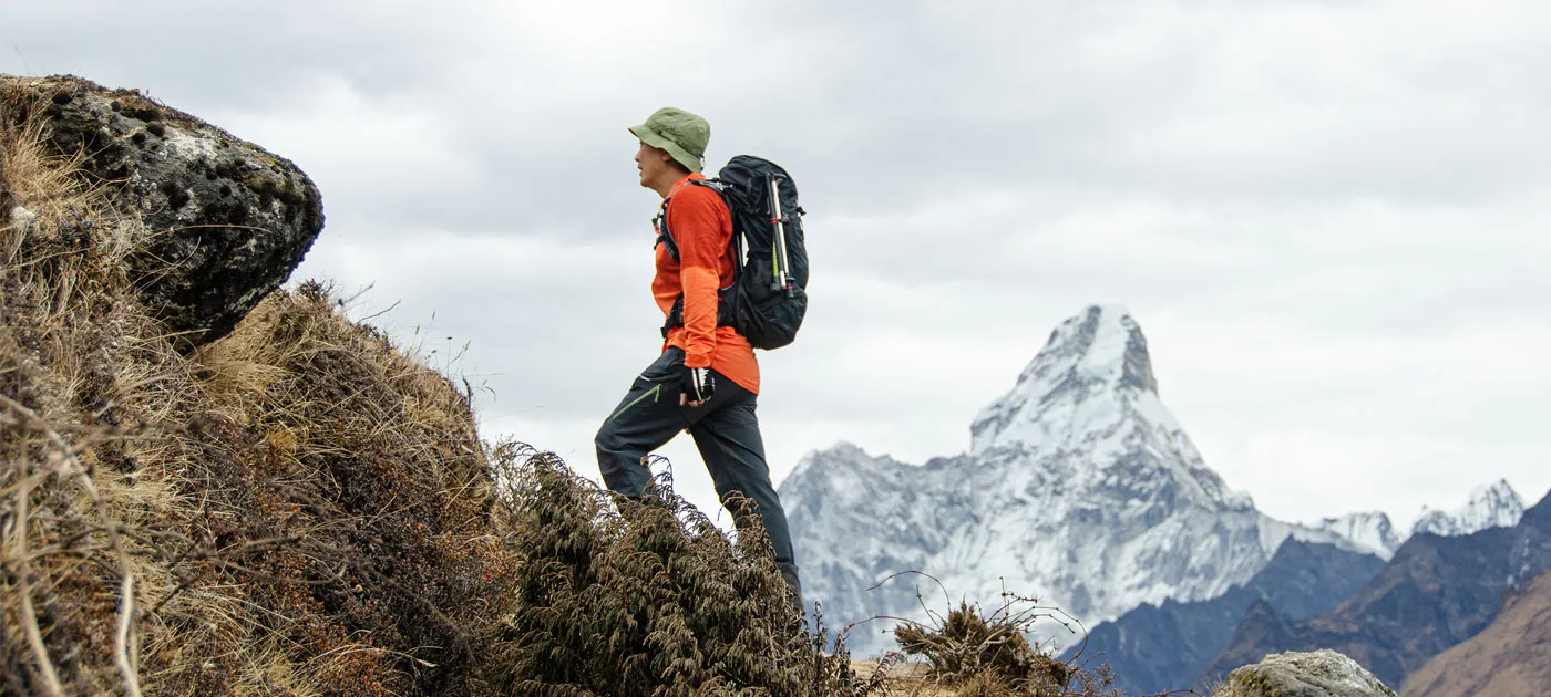 A male trekker in an orange long-sleeve shirt and sun hat walking uphill on a steep trail with Mount Ama Dablam visible in the background.
